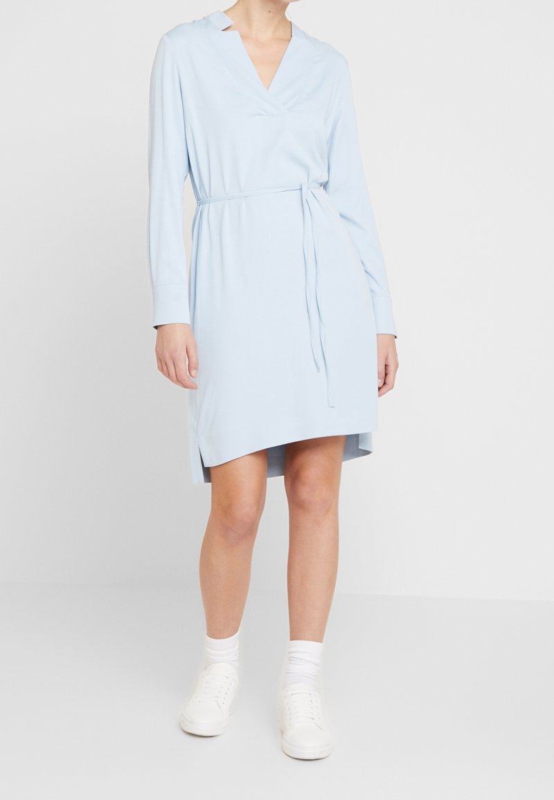 Vestido envolvente azul claro con mangas largas, escote en V, abertura lateral y cinturón tipo lazo. Combinado con zapatillas blancas y calcetines hasta el tobillo. Textura de tela suave.