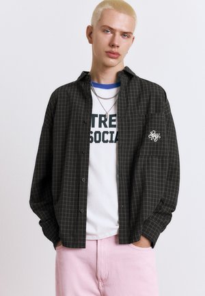 Mladá osoba s krátkymi blond vlasmi má na sebe viacvrstvové biele tričko s potlačou, čiernu kockovanú košeľu na gombíky, ružové nohavice a strieborné náhrdelníky.