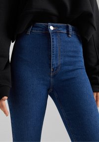 Jeans de talle alto azules hechos de denim, con cierre de cremallera, botones y patrones de costura visibles a lo largo de las costuras.