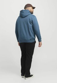 Blauwe hooded sweatshirt met een kangoeroezak, gecombineerd met zwarte broek en zwarte sneakers. Het model wordt van achteren gezien.