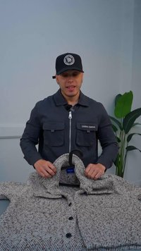 Grå stickad jacka med ett strukturerat mönster, mörka knappar och krage, hängande från en svart galge. Grön växt i bakgrunden.