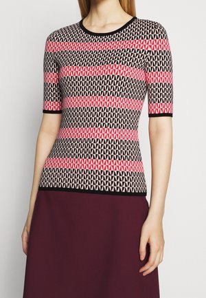 Femme portant un haut à manches courtes avec des motifs triangulaires noirs, blancs et roses, associé à une jupe violet foncé sur un fond uni.