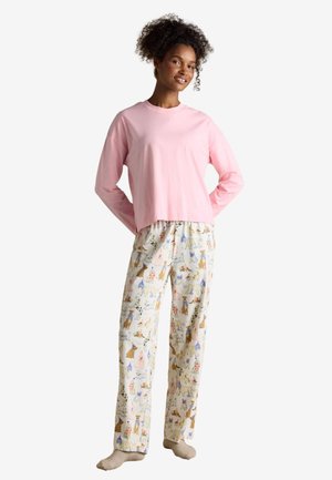 Kobieta stojąca z rękami za plecami, ubrana w jasnoróżową bluzkę z długim rękawem i białe spodnie piżamowe z kolorowymi ilustracjami psów.