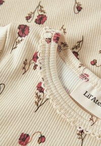 Haut en coton côtelé crème avec des imprimés floraux rouges, encolure festonnée et petite étiquette logo, texture douce et coupe ajustée.
