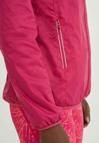 Gros plan sur le bras d'une personne portant une veste rose vif avec une poche zippée et des leggings roses à motifs.