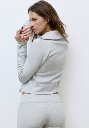 Femeie în hanorac gri deschis și pantaloni sport întinzându-și brațele pe un fundal alb simplu, privind concentrată.