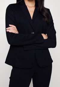 Donna con lunghi capelli scuri, che indossa un blazer nero aderente e pantaloni neri abbinati, con le braccia incrociate, su uno sfondo chiaro e semplice.