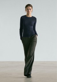 Marineblaues Langarmoberteil kombiniert mit dunkler, vertikal gestreifter, weiter Hose. Das Model steht selbstbewusst mit den Händen in den Taschen vor einem neutralen Hintergrund.