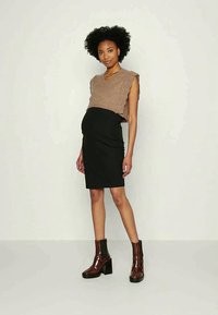 Mulher grávida vestindo um suéter sem mangas bege, saia preta até o joelho e botas de tornozelo castanhas, em pé contra um fundo claro e simples.