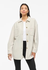 Ανοιχτό μπεζ oversized μπουφάν με δύο μπροστινές τσέπες, κούμπωμα με κουμπιά και κοφτό γιακά, φτιαγμένο από ύφασμα που μοιάζει με μαλλί.