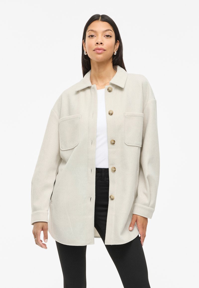 Ανοιχτό μπεζ oversized μπουφάν με δύο μπροστινές τσέπες, κούμπωμα με κουμπιά και κοφτό γιακά, φτιαγμένο από ύφασμα που μοιάζει με μαλλί.