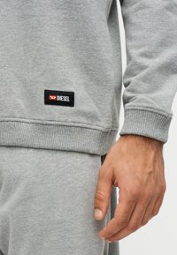 Κοντινό πλάνο ενός ατόμου που φοράει ανοιχτό γκρι φούτερ και παντελόνι με μαύρη ετικέτα Diesel στο φούτερ κοντά στη μέση.