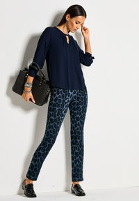 Vrouw in een marineblauwe blouse en blauwe broek met luipaardprint die een zwarte handtas vasthoudt, gekleed in zwarte loafers en dangling oorbellen, staand tegen een witte muur.