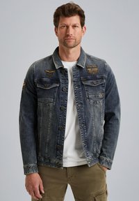 Veste en denim bleu foncé avec deux poches poitrine, fermeture à boutons et des patchs en cuir. Portée sur une chemise blanche à coupe décontractée.