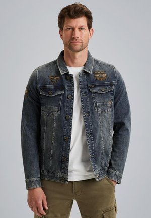 Denim jacket in dark blue with two chest pockets, button closure, and leather patches. Worn over a white shirt with a casual fit.