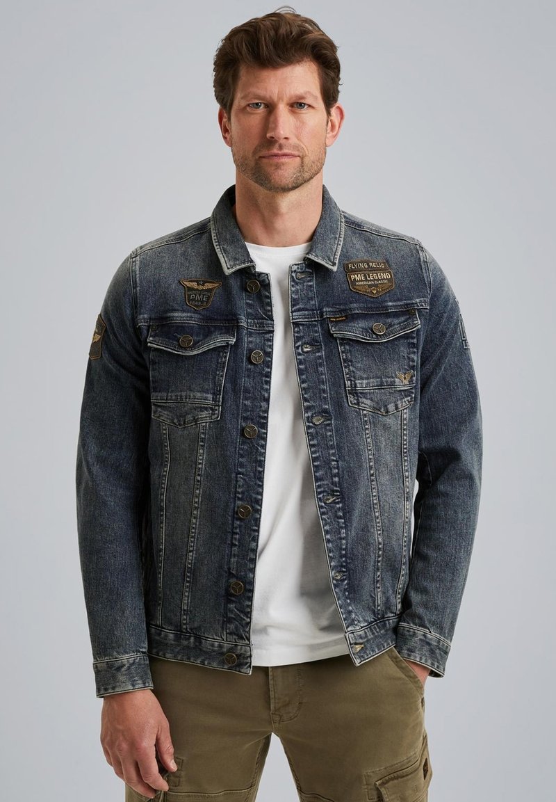 Veste en denim bleu foncé avec deux poches poitrine, fermeture à boutons et des patchs en cuir. Portée sur une chemise blanche à coupe décontractée.