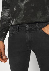 Černé džínové kalhoty se slim fit střihem, s matnou černou knoflíkem, pěti kapsami a jemnou strukturou. V kombinaci s tmavým trikem s dlouhým rukávem a tie-dye vzorem.