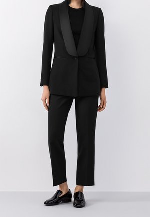 Femme portant un blazer noir ajusté avec des revers en satin, un pantalon noir assorti, un haut noir à manches longues et des mocassins noirs brillants.