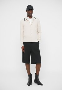 Beige kardigán fekete csíkkal ellátott gallérral, fehér inggel és fekete, térd felett érő szoknyával. Fekete bokacsizmával és fekete sapkával viselve.