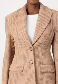 Béžový texturovaný blazer s pleteným vzorem, s klopami, dvěma předními kapsami a dvěma knoflíky. Tkanina vypadá lehce a prodyšně.