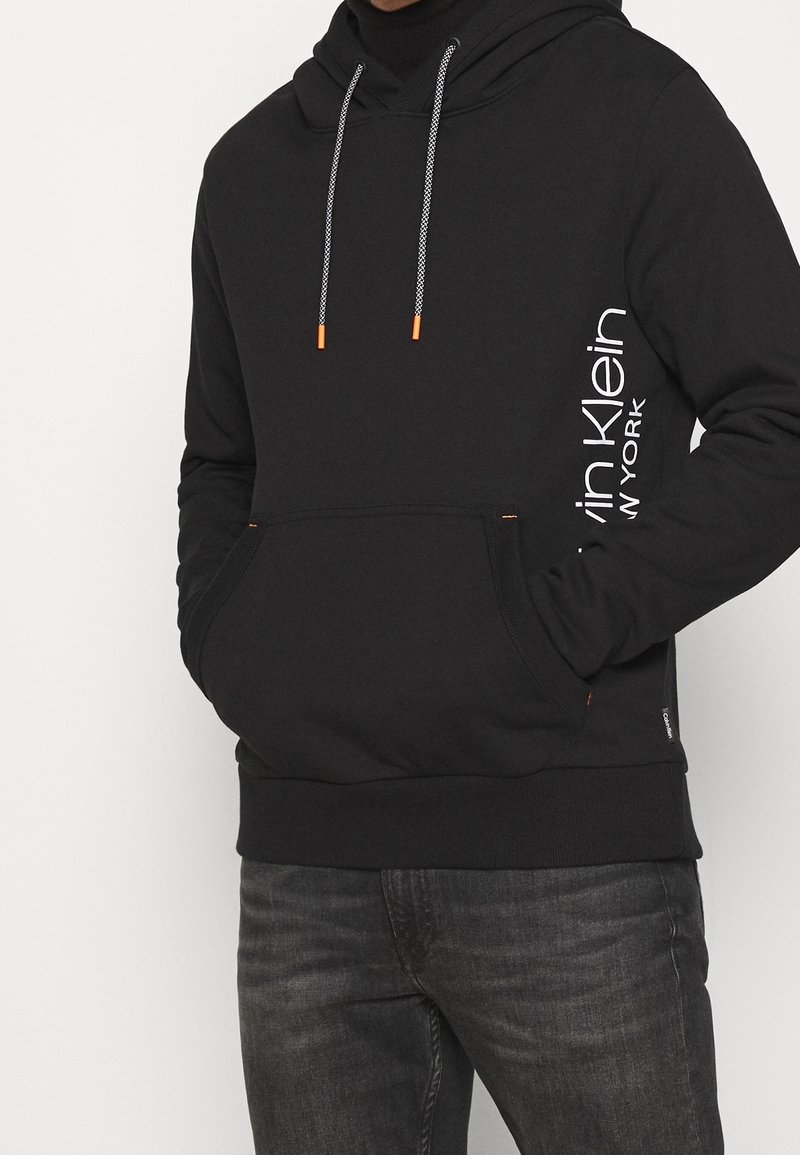 Sudadera negra con cordones, con puntas naranjas. Logo impreso verticalmente en un lado. Bolsillo tipo canguro y diseño de corte regular.