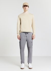 Beige Langarmshirt mit Rundhalsausschnitt, graue Cargo-Hosen mit Seitentaschen und weißen Sneakers. Die Kappe ist beige. Strukturierter Stoff.