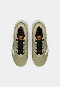 Beige un melnas Nike kedas ar melnām šņorēm, tenisa logotips uz mēles un mīksts iekšējais oderējums uz balta fona.