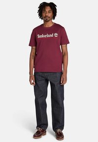 Vinröd bomullst-shirt med en Timberland-logga i vitt och guld, kombinerad med löst sittande mörka denimjeans och bruna skor.