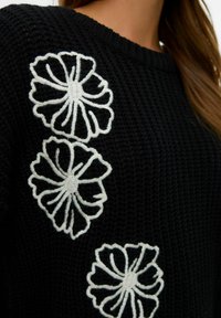 Zwart gebreid sweater met drie witte geborduurde bloemen aan de voorkant. Het ontwerp heeft een geribde halslijn en een textuurstof.