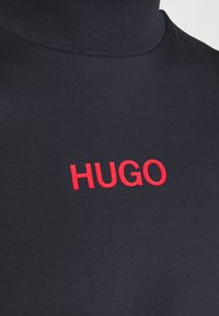 Marineblauer Rollkragenpullover mit glatter Textur und einem auffälligen roten "HUGO"-Logo, das auf der Vorderseite gestickt ist. Einfaches und modernes Design.