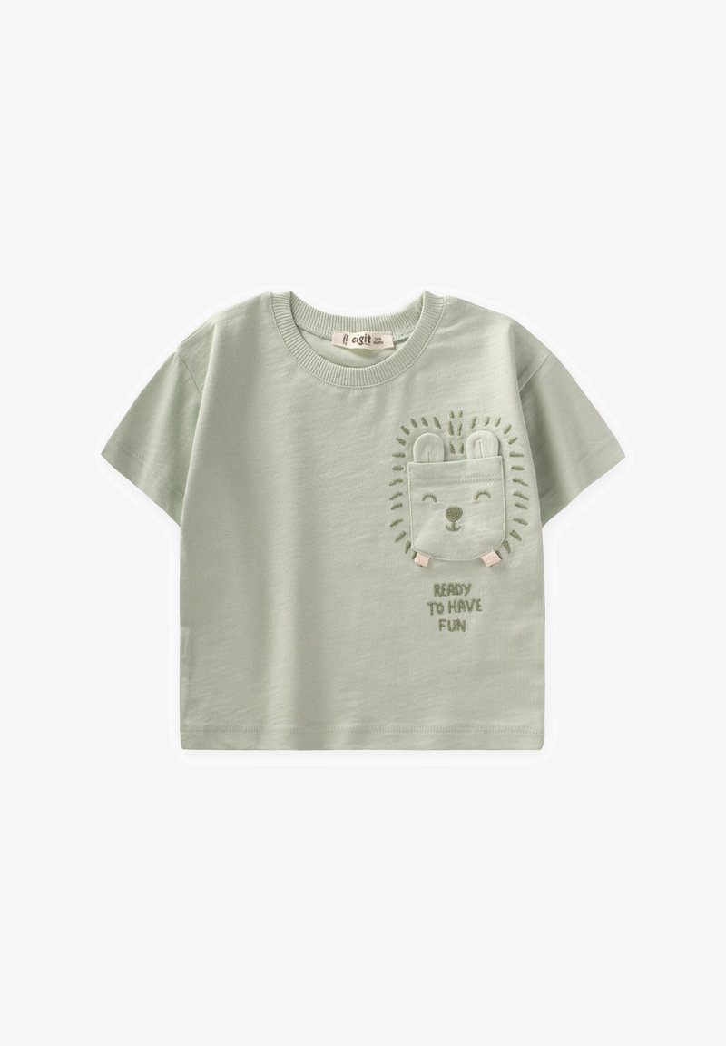 Gaiši zaļa kokvilnas T-krekls ar īsām piedurknēm, ar izšūtu lauvu sejas kabatu un tekstu "GATAVS IZKLAIDĒTIES" zemāk.