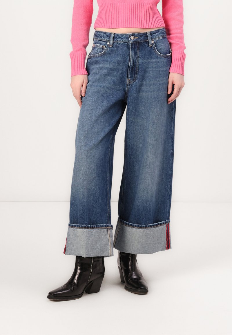 Jeans de pierna ancha en azul con puños de denim enrollados que presentan acentos en rojo y azul, combinados con elegantes botas negras de tacón y un suéter de punto rosa.