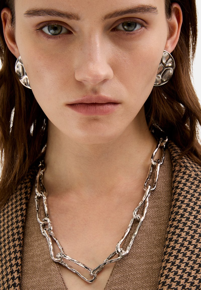 Sølv erklæringsohrhæng og en matchende tekstureret kæden Halskæde båret over en brun hundetand-mønstret top. Glatte og polerede overflader.