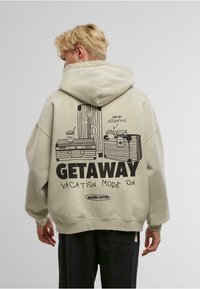 Hoodie oversize color beige chiaro con stampa grafica di valigie e una bevanda, con il testo "MODALITÀ VACANZA ATTIVATA" e una vestibilità rilassata.