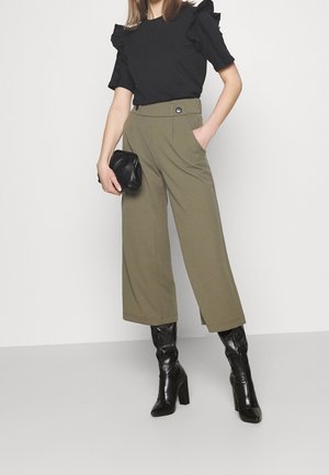 Femme en haut noir à volants à manches courtes, pantalon large cropped taille haute vert olive, bottes à talons noires, tenant une pochette noire.