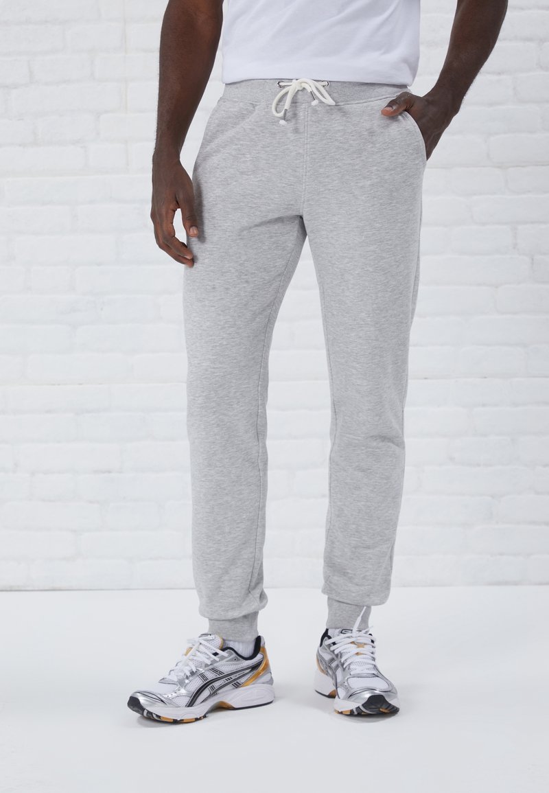 Pantalones de chándal grises con cintura ajustable, puños acanalados y bolsillos laterales. Combinados con zapatos deportivos plateados y negros. Diseño simple.