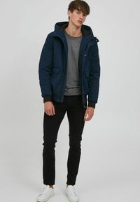 Navy hooded jacket with a zippered front, two front pockets, and ribbed cuffs, paired with a gray shirt and black pants. White sneakers included.