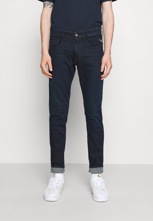 Mężczyzna ubrany w ciemnoniebieskie podwinięte dżinsy, białe trampki Adidas i granatową koszulę, stojący na jednolitym jasnym tle.