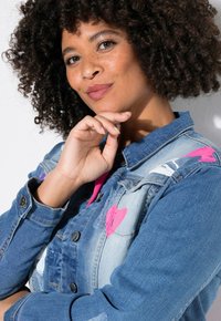 Chaqueta de denim en azul claro con acentos en forma de corazón rosa, detalles desgastados y botones de metal negro. Cuello clásico y mangas largas.