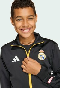 Musta urheilutakki keltaisella vetoketjulla, jossa on Real Madridin logo ja valkoiset Adidas-raidat. Pinta on teksturoitu ja istuu tiukasti.