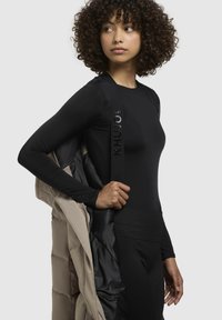 Camiseta negra de manga larga con textura suave y cuello redondo; chaqueta acolchada color beige parcialmente abierta, mostrando el forro negro. Cintura y pantalones negros.