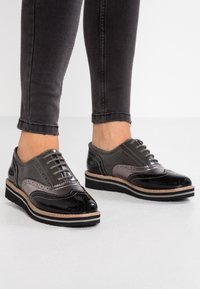 Chaussures en cuir grises et noires avec un design brogue, présentant un embout brillant, des accents texturés et une semelle à plusieurs couches avec un bord marron clair.