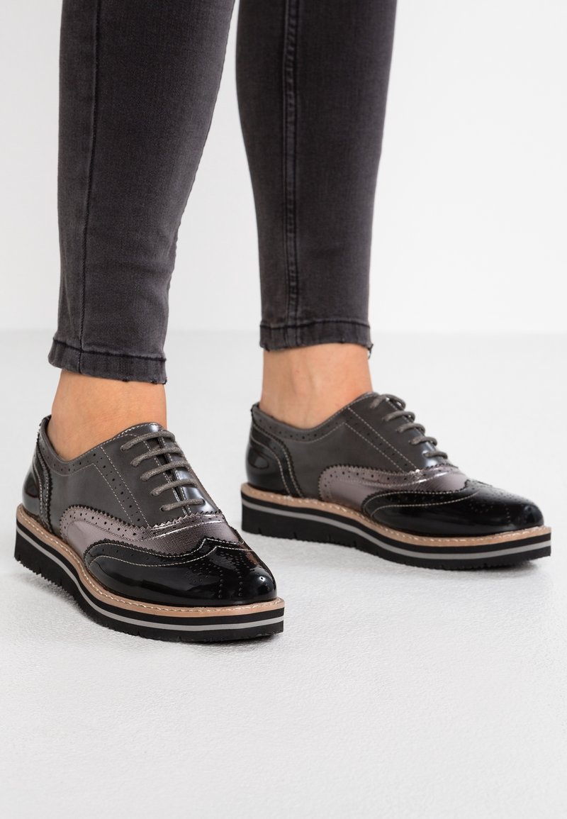 Chaussures en cuir grises et noires avec un design brogue, présentant un embout brillant, des accents texturés et une semelle à plusieurs couches avec un bord marron clair.