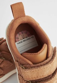Brązowy sneaker z teksturowaną górną częścią z tkaniny, skórzanymi akcentami i paskiem na rzep. W środku znajduje się etykieta z napisem "TO NALEŻY DO".