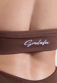 Sujetador deportivo marrón acanalado con una banda elástica ancha que presenta un logotipo bordado en blanco. Textura suave y mate sobre la piel visible en el fondo.