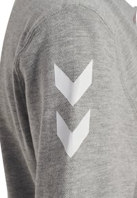Grå sweater med tekstureret stof og hvide chevron-patches på ærmerne. Detaljeorienteret design med et enkelt, moderne udtryk.
