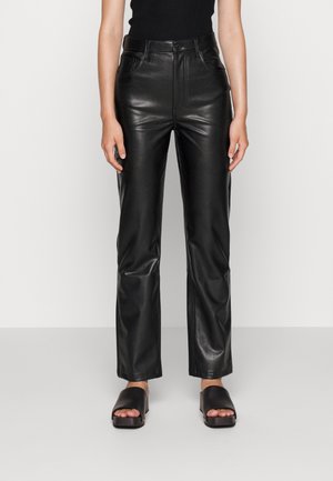 Person trägt schwarze Lederhose mit geradem Bein und dazu schwarze offene Sandalen, steht vor einem einfarbigen hellen Hintergrund.