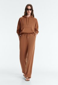 Femme portant un sweat à capuche marron et un pantalon large assorti, des lunettes de soleil et des sandales noires, marchant les mains dans les poches sur fond uni.