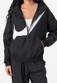 Svart lättviktsjacka med dragkedja, vit logotyp och huva. Har elastiska muddar och fåll. Materialet ser slätt ut med en avslappnad passform.