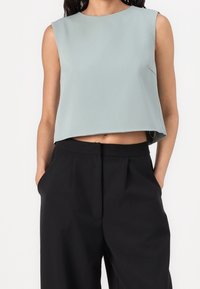 Donna che indossa un top azzurro chiaro senza maniche, corto, abbinato a pantaloni neri larghi a vita alta, con le mani nelle tasche.